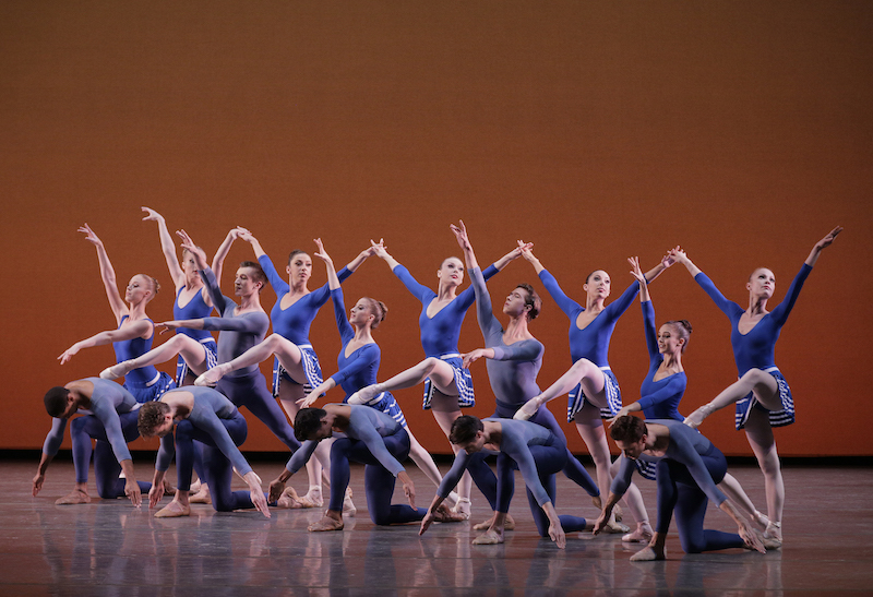 A group of dancers in blue costumes, evocative of Coco Chanel 1920s bathing suits, stand in three long lines. Some kneel, others lunge and a few rest their raised legs on the backs of others.